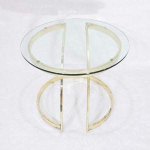May include: A round glass top coffee table with a gold metal frame. The frame is made of two large circles that intersect in the middle, creating a unique design.