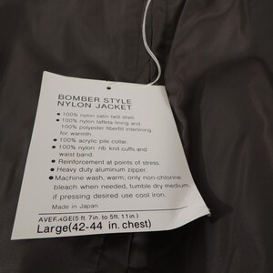 May include: A dark gray bomber style nylon jacket with a tag attached. The tag lists the jacket's features, including a nylon satin twill shell, acrylic pile collar, and heavy-duty aluminum zipper. The jacket is a Large size, with a chest measurement of 42-44 inches.