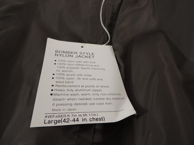 May include: A dark gray bomber style nylon jacket with a tag attached. The tag lists the jacket's features, including a nylon satin twill shell, acrylic pile collar, and heavy-duty aluminum zipper. The jacket is a Large size, with a chest measurement of 42-44 inches.