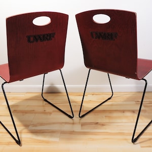 Puede incluir: Dos sillas de madera roja con patas de metal negro. Las sillas tienen un recorte en el respaldo y las palabras "UWRF" están impresas en la parte posterior.