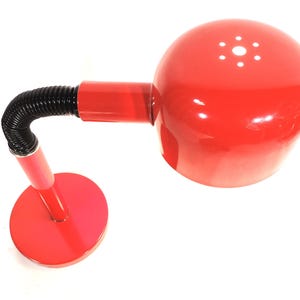 Op de afbeelding: Rode bureaulamp met een ronde voet en een flexibele zwarte nek. De lamp heeft een bolvormige kop met kleine gaatjes. De lamp is gemaakt van metaal en plastic.