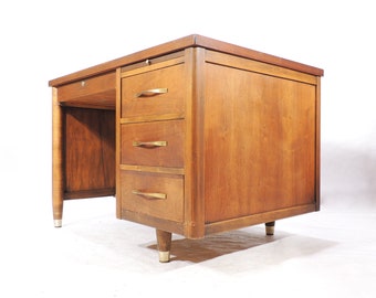May include: A mid-century modern desk with a wood grain finish and brass hardware. The desk has three drawers and a single drawer on the left side. The desk has tapered legs.