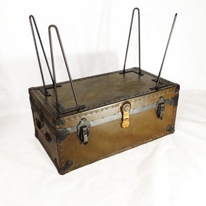 May include: A vintage green metal trunk with black metal hardware and a brass lock. The trunk is sitting on four metal legs.