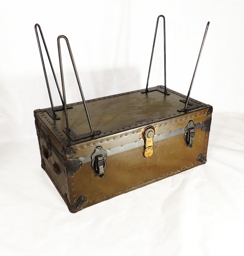 May include: A vintage green metal trunk with black metal hardware and a brass lock. The trunk is sitting on four metal legs.