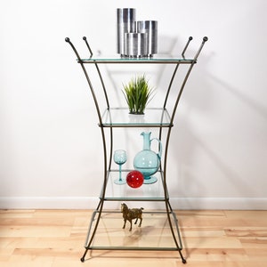Puede incluir: Una estantería de cuatro niveles de vidrio y metal con un diseño curvo. Las estanterías son de vidrio transparente y el marco es de color bronce oscuro. La estantería superior tiene tres jarrones cilíndricos plateados, la segunda estantería tiene una pequeña planta en maceta, la tercera estantería tiene una jarra de vidrio azul, una copa de vino transparente y una bola de vidrio roja, y la estantería inferior tiene una pequeña figura de caballo de metal dorado.