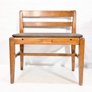 Puede incluir: Una silla de madera con acabado marrón claro. La silla tiene un asiento rectangular cubierto con una tela gris. El respaldo presenta tres listones horizontales de madera. La silla tiene cuatro patas rectas y una barra de soporte horizontal.