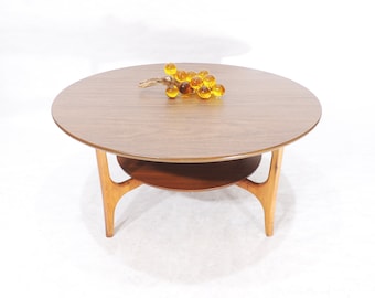 May include: A round wooden coffee table with a lower shelf. The table has a light brown finish and unique, curved legs. A cluster of amber glass spheres sits on the table top.