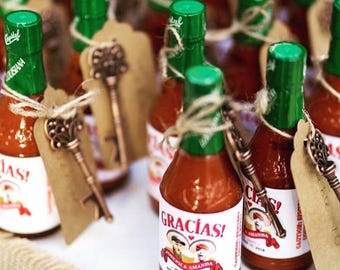 May include: Small glass bottles of hot sauce with green caps and white labels that read "Gracias!" and "Joseph & Amanda, Wed Hot! November 17, 2018." Each bottle is adorned with a decorative key and a tag.