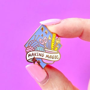 May include: A Making Magic enamel pin featuring crafting tools and yarn in shades of blue, pink, and yellow. The pin has a banner with the words "MAKING MAGIC" on it. The pin is held up against a purple background.