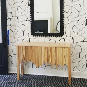 Puede incluir: Un espejo negro arqueado con un borde de cuentas cuelga sobre una mesa consola de madera con un borde inferior único y dentado. La mesa consola está frente a una pared blanca con un papel tapiz de patrón geométrico blanco y negro.
