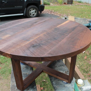 Op de afbeelding: Een ronde houten salontafel met een donkerbruine walnoot afwerking. De tafel heeft een eenvoudig, modern ontwerp met een kruisvormige basis.