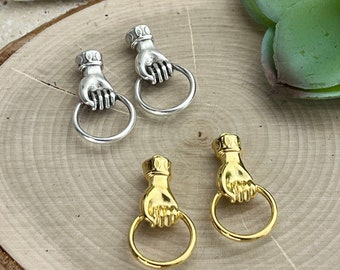 Könnte beinhalten: Zwei Paar Ohrringe, ein Paar ist silber und das andere ist gold. Beide Paare zeigen eine Hand, die einen Ring hält. Die Ohrringe liegen auf einer Holzoberfläche.