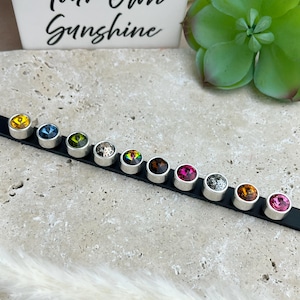Peut inclure: Un bracelet en cuir noir avec une rangée de strass en cristal colorés. Les strass sont sertis dans des montures métalliques argentées. Le bracelet est sur une surface grise.