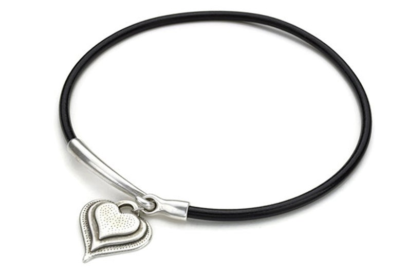 May include: A black leather bracelet with a silver heart charm. The heart charm has a textured surface and a double layered design.