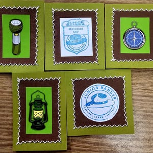 May include: Five square cards with a green background and brown border, each featuring a different outdoor-themed graphic. Images include a flashlight, compass, lantern, and Junior Ranger badges. White stitching frames each card.