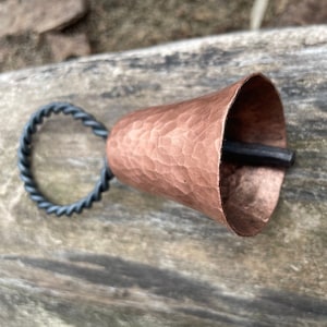May include: A hammered copper bell with a twisted black metal handle. The bell has a rustic, handmade look.