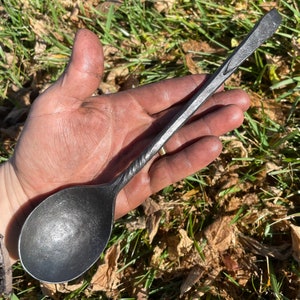 Könnte beinhalten: Eine Hand hält einen schwarzen Löffel aus Metall mit einem gedrehten Griff. Der Löffel ist aus geschmiedetem Stahl und hat eine glatte, abgerundete Schale.