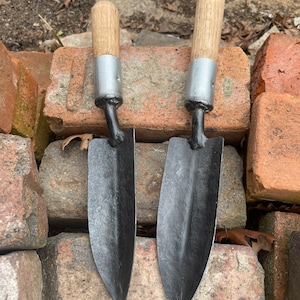 Peut inclure: Deux outils de jardinage à manche en bois et lames en métal noir. Les outils sont posés sur une pile de briques rouges.