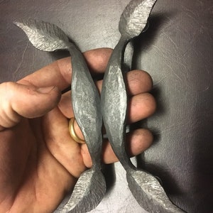 May include: Two hand-forged metal door pulls shaped like leaves. The pulls are a dark gray color and have a rustic finish.