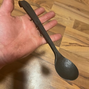 Puede incluir: Un cucharón de metal negro con un mango largo. El mango tiene una forma de gancho única en la parte superior.