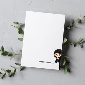 May include: A white card with a black cartoon ninja peeking out from the right side. The card has the text "PSSSSSST...." printed on it.