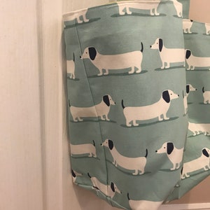 May include: A light green canvas tote bag with a white dachshund pattern. The bag has two handles and is made of a durable fabric.