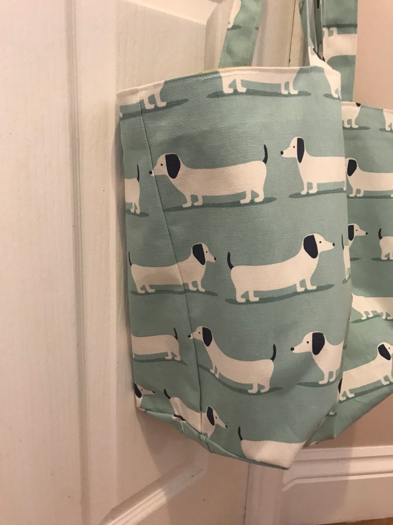 May include: A light green canvas tote bag with a white dachshund pattern. The bag has two handles and is made of a durable fabric.