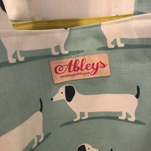 May include: A green canvas tote bag with a white dachshund pattern. The bag has a yellow fabric lining and a brown leather tag that says "Abbley's Handmade With Love".