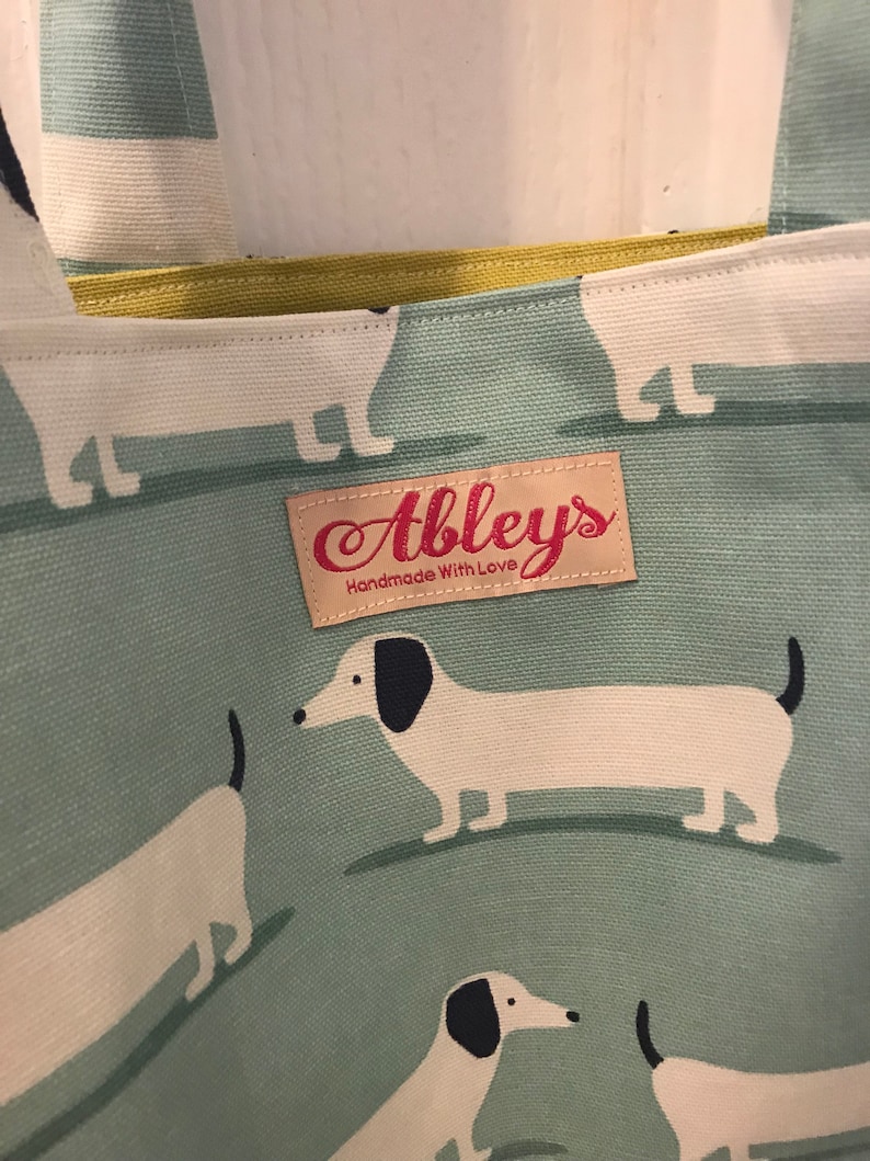 May include: A green canvas tote bag with a white dachshund pattern. The bag has a yellow fabric lining and a brown leather tag that says "Abbley's Handmade With Love".