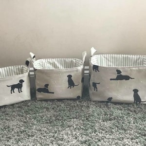 May include: Three fabric storage baskets with black labrador retriever silhouettes on a white background. The baskets are lined with a gray and white striped fabric. The baskets are different sizes.
