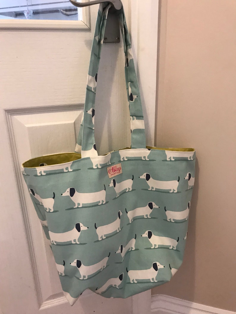 May include: A light blue tote bag with a white and black dachshund pattern. The bag has a yellow lining and a small white label with the text "Abigael" on it.