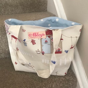 May include: A white tote bag with a blue and white polka dot lining. The bag features a red and white beach hut design with the text "Abbleys Handmade With Love".