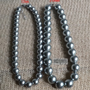 Peut inclure: Deux colliers de perles grises, l'un avec des perles de 12 mm et l'autre avec des perles de 16 mm. Les colliers sont disposés sur un fond en tissu beige.