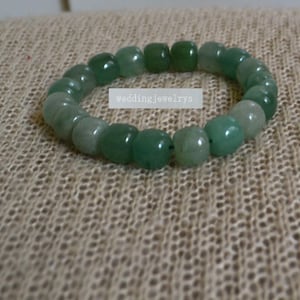 Könnte beinhalten: Ein Armband aus grünen Edelsteinen, das aus runden Perlen besteht. Das Armband liegt auf einem hellbraunen Stoffhintergrund.