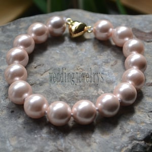 Puede incluir: Un brazalete hecho de perlas de color rosa claro con un cierre de corazón dorado. El brazalete está sobre una superficie gris texturizada.
