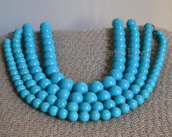 May include: A turquoise statement necklace with multiple strands of round beads. The necklace has varying bead sizes, creating a layered effect. The beads are a vibrant turquoise colour, and the necklace is displayed on a neutral surface.