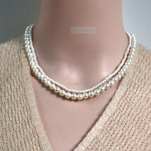 Puede incluir: Un collar de perlas de doble cadena con perlas blancas. El collar está en un maniquí blanco que lleva un suéter beige.