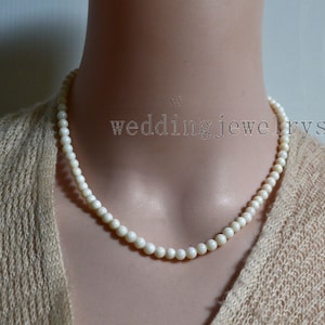 May include: A cream-coloured beaded necklace with the text "wedding jewelrys" in a light grey font. The necklace is draped around a person's neck, and the beads appear to be small and round. The background is a neutral colour.
