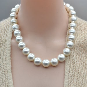 Op de afbeelding: Een klassieke witte parelketting met grote, ronde parels. De ketting is gemaakt van een rij parels die gelijkmatig verdeeld zijn en met een knoop tussen elke parel aan elkaar zijn gebonden.