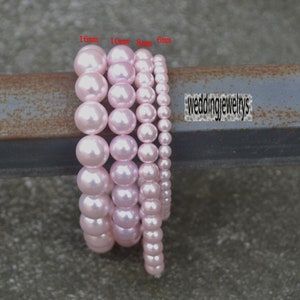 Puede incluir: Cuatro hebras de cuentas de perlas rosas de diferentes tamaños, 16 mm, 10 mm, 8 mm y 6 mm, se muestran sobre una superficie metálica. El texto "weddingjewelrys" es visible en el fondo.