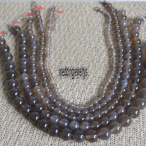 May include: Five strands of grey agate beads in various sizes, ranging from 6mm to 14mm. The beads are round and smooth, with a slight sheen. The strands are laid out on a textured surface, with the sizes labelled in millimetres.