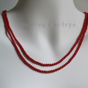 Può includere: Collana di perline rosse a due fili. La collana è composta da piccole perline rotonde ed è esposta su un manichino bianco. Il testo "wedding jewelrys" è debolmente visibile sullo sfondo.