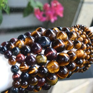 May include: Beaded bracelets made from polished tiger eye stones. The beads display a range of colours, including brown, gold, and blue. The bracelets are arranged in a layered design, showcasing the natural patterns of the stones. The beads vary in size.
