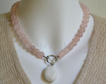 Puede incluir: Collar de cuentas de cuarzo rosa con un colgante ovalado blanco. El collar tiene un cierre plateado y se muestra en un maniquí. Las cuentas son de color rosa pálido.