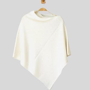 Könnte beinhalten: Ein weißer Strickponcho mit einem weiten Ausschnitt. Der Poncho hängt an einem Holzbügel.