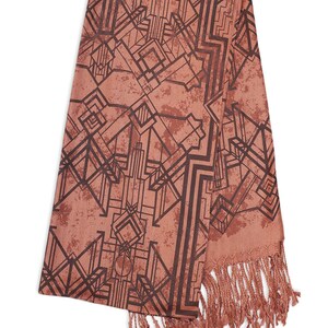 May include: A coral-colored scarf with a geometric Art Deco design in dark brown. The scarf has a fringed edge and a textured appearance. The pattern features angular shapes and lines, creating a vintage aesthetic.