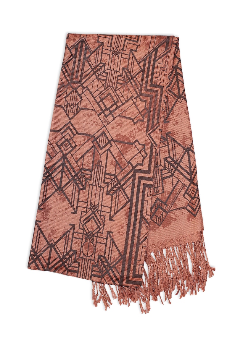 May include: A coral-colored scarf with a geometric Art Deco design in dark brown. The scarf has a fringed edge and a textured appearance. The pattern features angular shapes and lines, creating a vintage aesthetic.