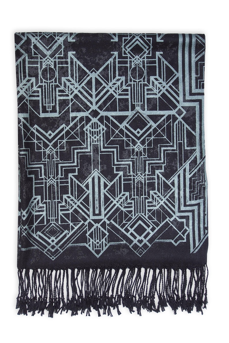 May include: A black scarf with a geometric Art Deco design in light blue. The scarf has a fringe along the bottom edge. The pattern features lines, shapes, and repeating motifs.