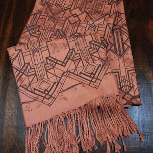 May include: A terracotta-colored scarf with a geometric art deco design in dark brown. The scarf has a fringed edge. The scarf is made of a soft material and is suitable for wear in the fall and winter.