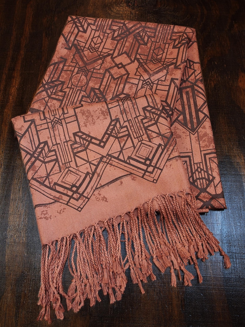 May include: A terracotta-colored scarf with a geometric art deco design in dark brown. The scarf has a fringed edge. The scarf is made of a soft material and is suitable for wear in the fall and winter.